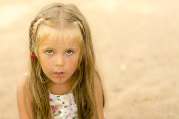 Little dirty blond girl with a pigtail gazes ahead on the background of sand