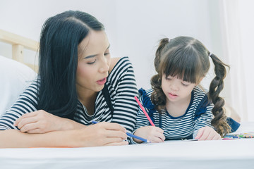 A little cute girl enjoy drawing into the book by colorful pastel with mother on the white bed in the bedroom, education time at home