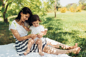 Pretty mother and her little daughter playing outdoors, explore green grass. Cute mom and her cheerful child playing in the park together. Portrait of happy family. Motherhood and childhood.
