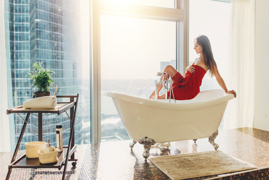Slim young woman in towel sitting on bathtub in luxurious bathroom