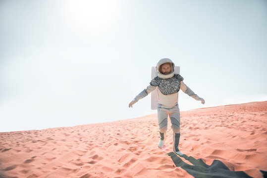 Serious spaceman wearing white armor is hardly trying to move forward on desert and looking at camera with determination. Full length portrait. Copy space on left side