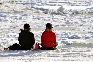 Fototapeta na wymiar Rear view of couple sitting on the snowy beach and looking at the frozen sea.