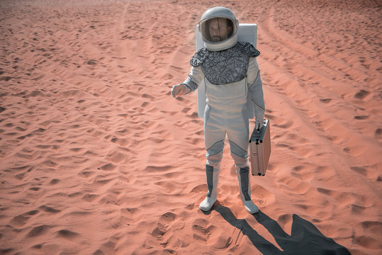 Serious spaceman wearing white armor is standing on red sand under hot sun and rising hand. He holding small suitcase. Full length portrait. Copy space on left side