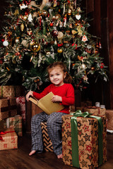 little girl is reading stories in a book by a Christmas tree. Red shirt of pajamas. Sits on gift boxes.