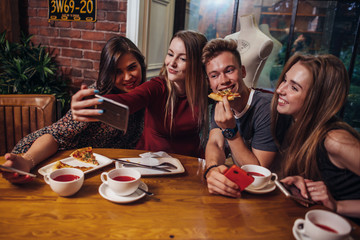 Group of cheerful young friends taking selfie with smartphone having light dinner in modern stylish restaurant