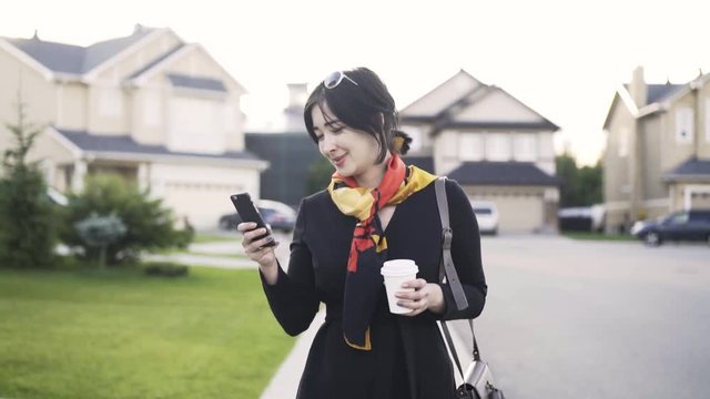 Cheerful and beautiful Asian bissinesswoman with a coffee to go is looking at her phone and walking in a suburbian area. Tracking real time establishing shot