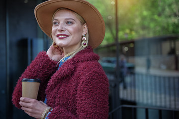 Waist up portrait of dreamful young woman enjoying hot beverage outdoor. She is looking aide...