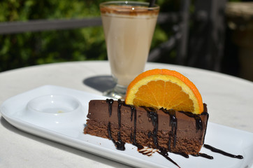 Chocolate cake with rich topping and a slice of orange - on white porcelain plate - and a glass of cream coffee or a macchiato