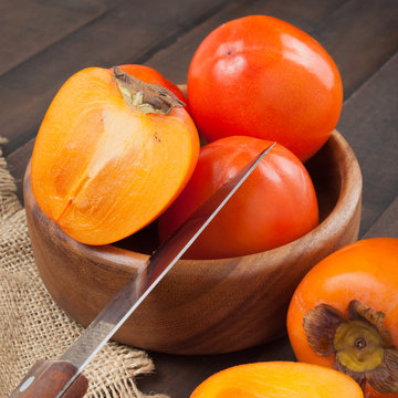 Persimmons in bowl with kitchen knife on wooden table.