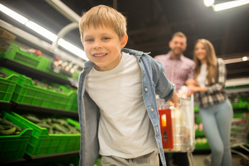 Portrait of happy little boy having fun in supermarket buying groceries with family, pulling shopping cart behind him and smiling cheerfully at camera