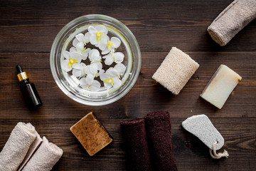 Bath in bowl with tropical flowers for foot spa, pumice stone, soap and oil on dark wooden background top view