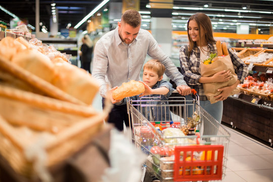 Portrait of happy young family  shopping for groceries in supermarket together with little boy, while choosing fresh bread loaf in bakery department