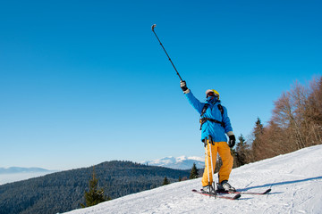 Full length shot of a professional skier taking a selfie using selfie stick posing on the slope. Blue sky, mountains and winter forest on the background