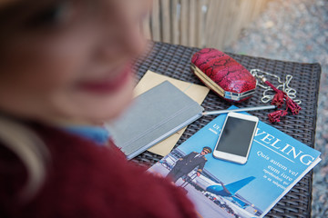 Top view close up of cheerful woman smiling while sitting outdoor. Focus on magazine about traveling, smartphone, notebook and small cutch bag on table