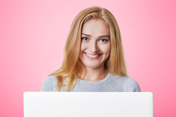 Naklejka premium Cheerful female model with happy expression smiles gladfully, uses laptop computer, surf social networks and recieves messages from friends, uses wireless internet connection. Technology concept