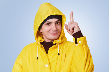 Clever handsome man raises fore finger as gets brilliant idea, wears yellow raincoat, has happy...