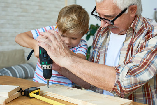 Portrait of senior man helping little boy make wooden model, teaching him carpentry and drilling boards