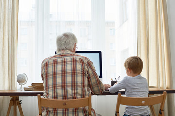 Rear view portrait of grandfather and grandson doing homework together sitting at desk with  blank screen computer