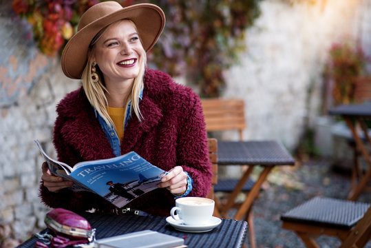 Portrait of excited young woman reading journal about traveling with enjoyment. She is laughing while having rest in cozy cafeteria. Copy space