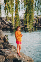 Outdoor portrait of cute preteen kid girl playing by the lake at sunset, wearing yellow pullover and red shirts