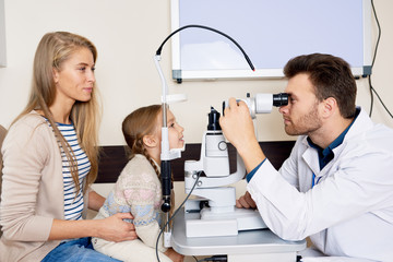 Side view portrait of young optometrist testing sight of little girl using slit lamp during medical...