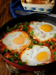 A pan of fried eggs with tomato sauce and parsley on a wooden background. Shakshuka a traditional meal of the Jewish cuisine