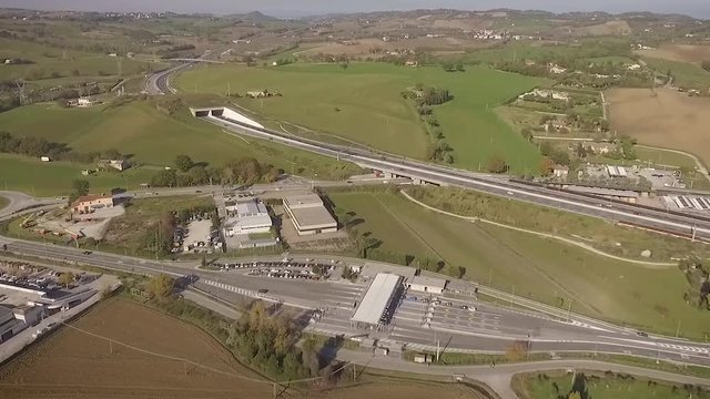 Vista aerea di un casello autostradale con traffico di auto e camion regolari. L'autostrada si trova in zona collinare