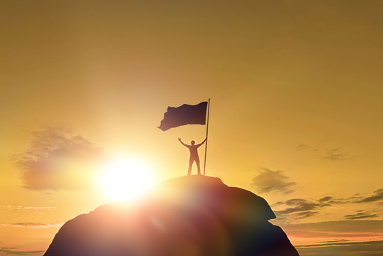 High achievement, silhouettes of men, victory flag on top of the mountain, hands up. A man on top of a mountain. Conceptual design. Against the dramatic sky with clouds at sunset.