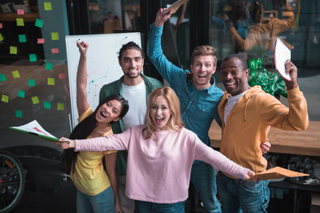 Fototapeta premium Best team ever. Top view portrait of extremely happy multinational coworkers are standing in office and screaming while expressing gladness. They are holding folders and looking at camera with joy