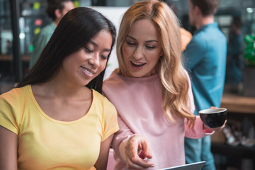 Do this way. Qualified attractive emotional girls are using laptop, looking at screen with smile while pointing on monitor and discussing business project. Attentive men are working in background