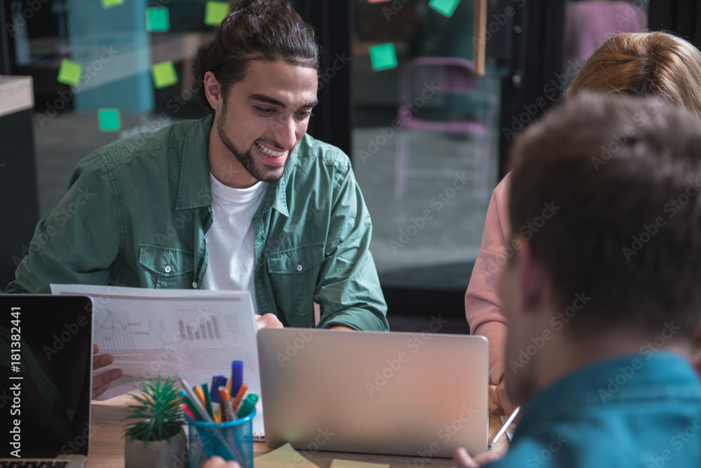 Poster portrait of beaming unshaven man watching at laptop while keeping sheet of paper with statistic in h