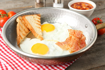 Frying pan with eggs, toasts and bacon on table