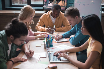 Obraz premium Orderly men and women locating at desk. Male making notes in copybook while female typing on laptop. Portrait of comrade situating near them. Occupation concept
