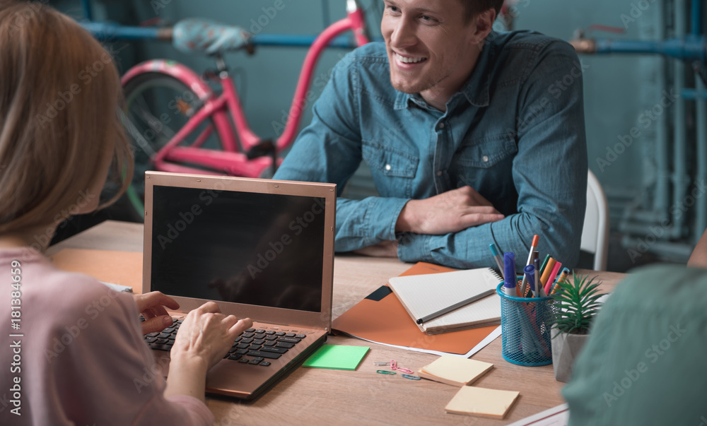 Poster outgoing man telling with female writing in notebook computer while locating at desk in room. job co