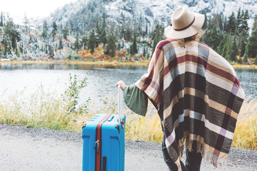 Woman wearing poncho and hat with bright blue suitcase looking at stunning mountain lake...