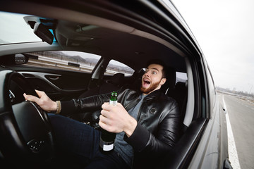 Drunk young man with a beard with a bottle of beer in his hand behind the wheel of a car. Emergency situation, violation of law, drunk driving