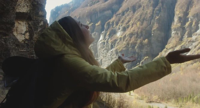 Caucasian female hiker in yellow raincoat wearing backpack standing under small waterfall, trying to catch drops with her hands. 4K UHD RAW edited footage