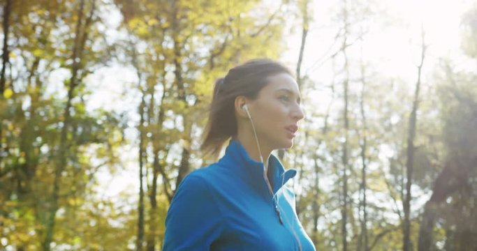 Close up from the side of young sporty pretty woman in blue sweater and with headphones jogging in the park. The sunny weather of early autumn. Outdoor. Slow motion