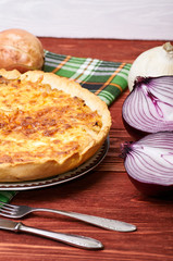 Appetizing French onion pie. A beautiful homemade pie with cheese crust stands on a green checkered napkin. Onions, cheese and cutlery on a rough wooden rustic table. Different kinds of onions.