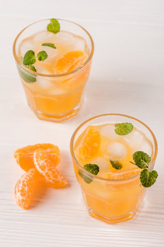 Mandarin cocktail with ice and mint in beautiful glasses and jug, fresh ripe citrus on white wooden background. Sweet orange juice. Close up photography. Selective focus
