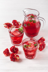 Pomegranate cocktail with ice and mint in beautiful glasses and jug, fresh ripe pomegranate on white wooden background. Sweet red juice. Close up photography. Selective focus