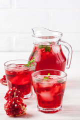 Pomegranate cocktail with ice and mint in beautiful glasses and jug, fresh ripe pomegranate on white wooden background. Sweet red juice. Close up photography. Selective focus