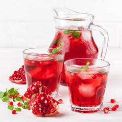 Fototapeta na wymiar Pomegranate cocktail with ice and mint in beautiful glasses and jug, fresh ripe pomegranate on white wooden background. Sweet red juice. Close up photography. Selective focus