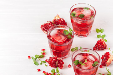 Pomegranate cocktail with ice and mint in beautiful glasses and jug, fresh ripe pomegranate on white wooden background. Sweet red juice. Close up photography. Selective focus