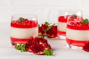 Creamy vanilla panna cotta with red jelly in beautiful glasses, fresh ripe pomegranate on white...