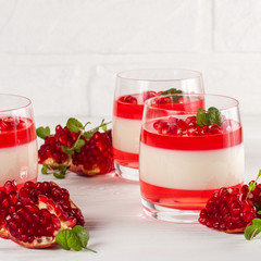 Creamy vanilla panna cotta with red jelly in beautiful glasses, fresh ripe pomegranate on white wooden background.