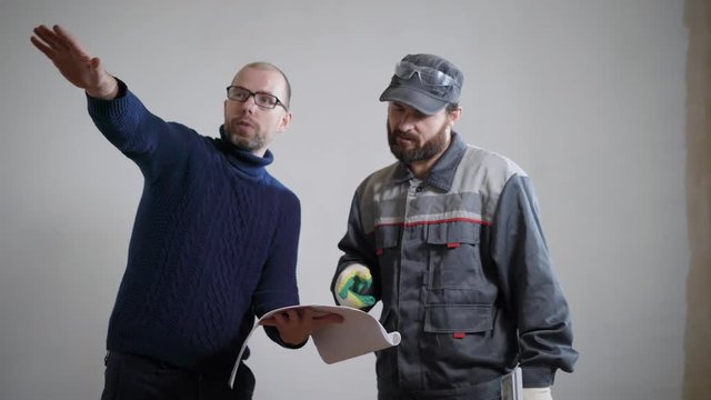 foreman is explaining for master of repair, holding project in hands, standing in a flat with empty white walls
