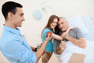 An elderly man lies in a hospital room on a bed. He is seen by a man with a woman. The woman is hugging the old man.