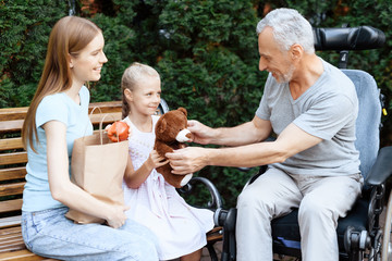 An elderly man is sitting in a wheelchair. He is seen by a woman with a girl. They are sitting on a bench in the park.