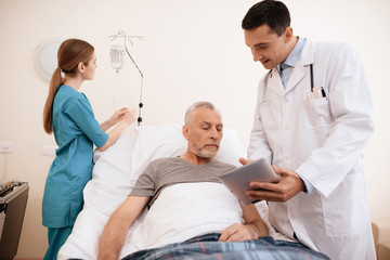 The old man lies on a cot in medical ward, and next to him stands a doctor. He is showing something to old man on tablet